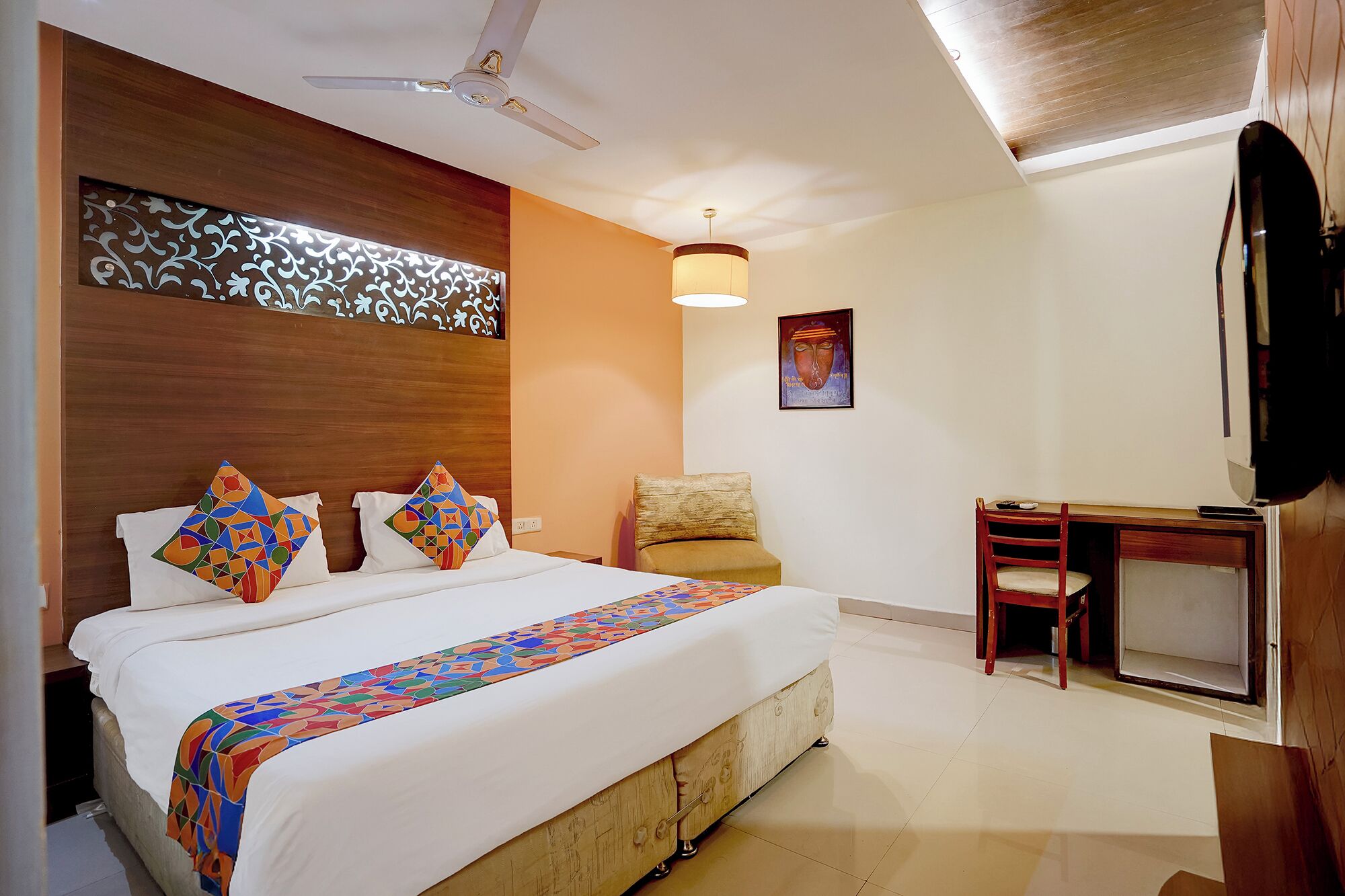 A hotel room with a bed, a desk, a chair, a ceiling fan, and a wall-mounted TV.