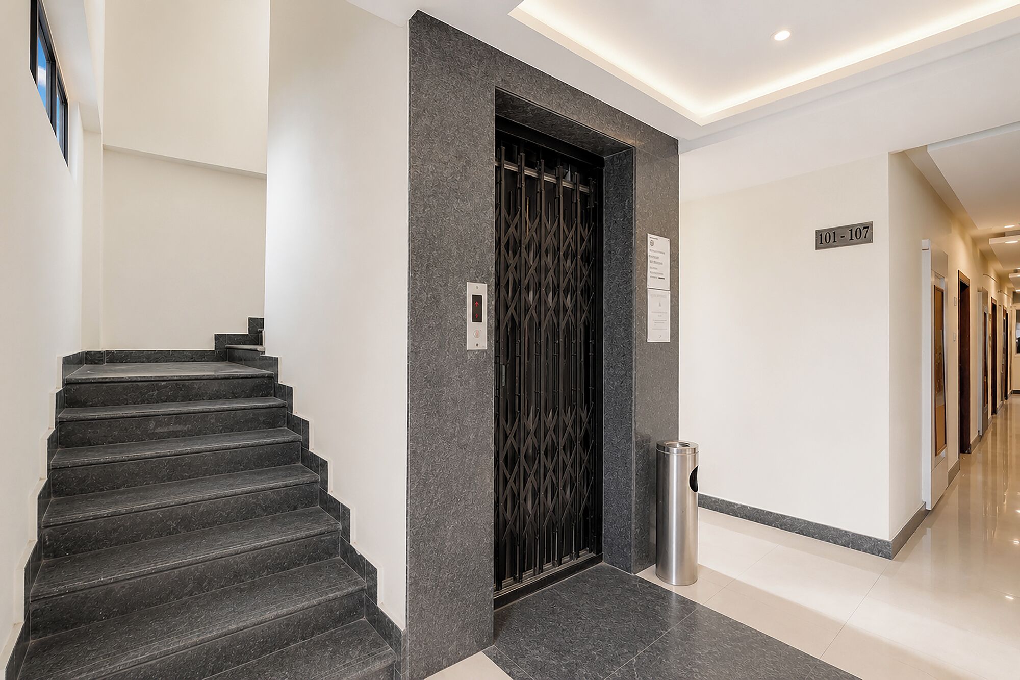 A modern hotel lobby with a staircase, a metal elevator, and a trash bin.