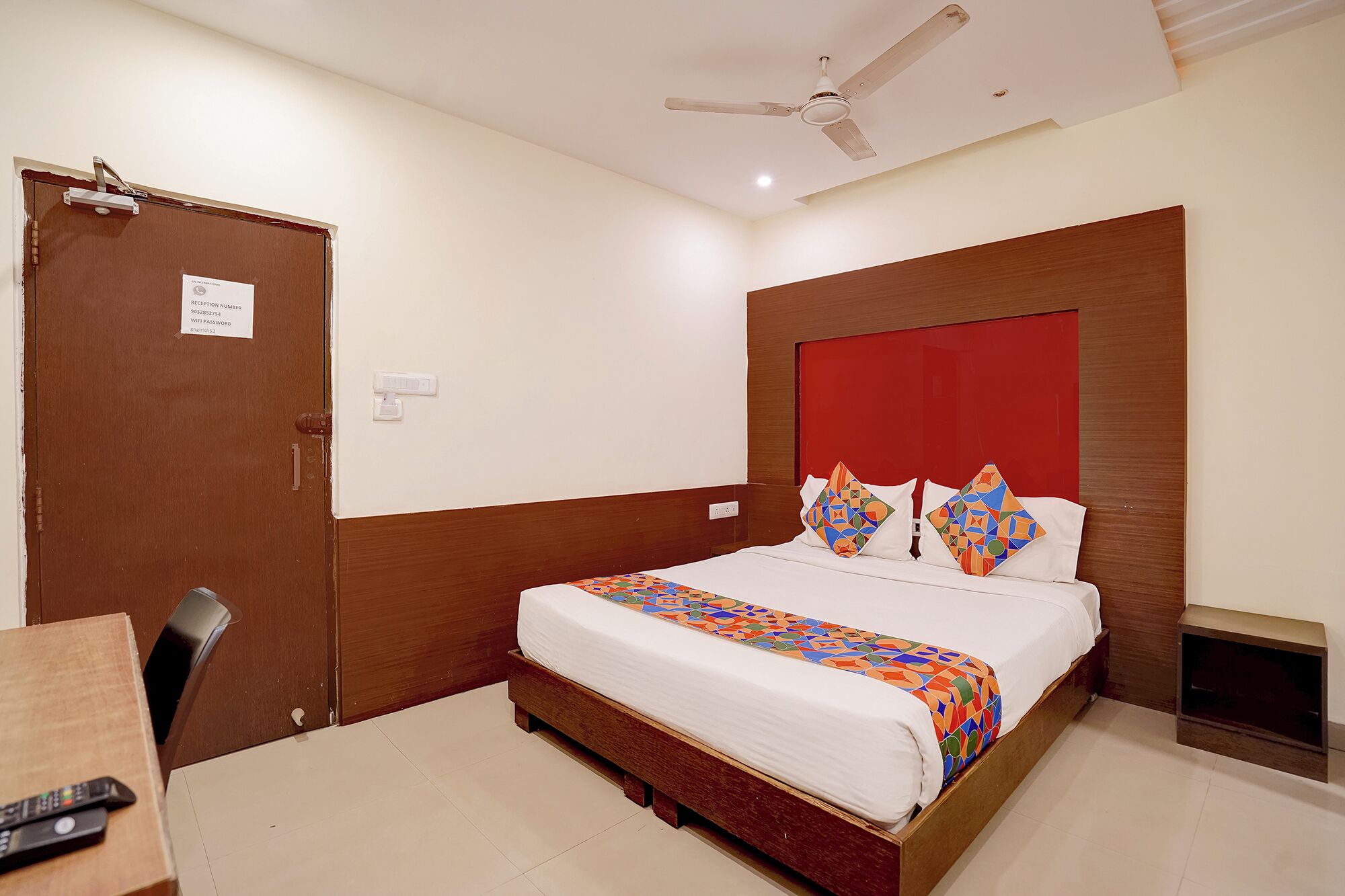 Habitación de hotel con cama, escritorio, silla, ventilador de techo y puerta con número de recepción.