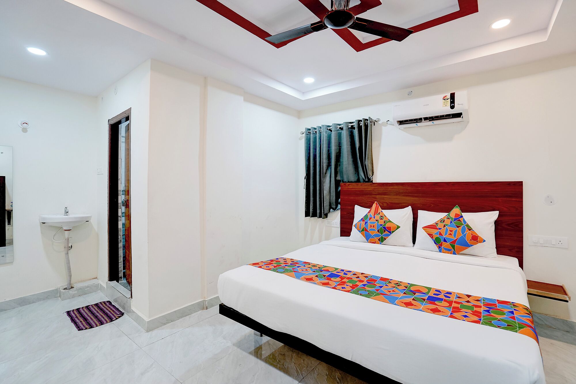 A hotel room with a bed, a ceiling fan, an air conditioning unit, a window with curtains, and a sink.
