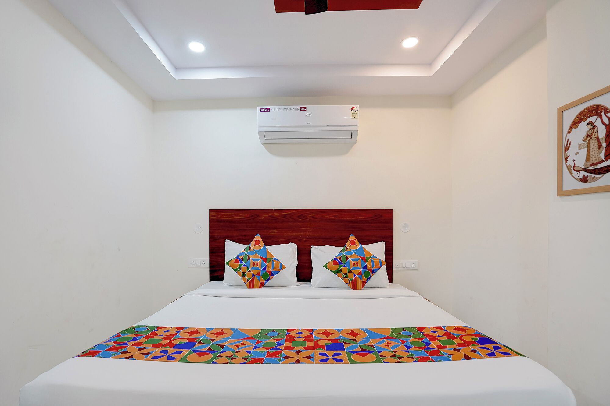 A hotel room with a bed, a ceiling light, an air conditioning unit, and a wall-mounted artwork.
