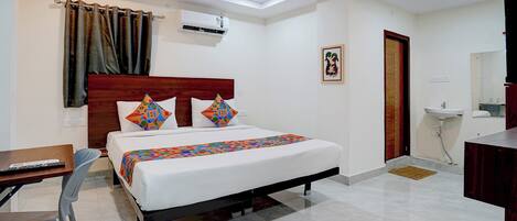 Habitación de hotel con cama, escritorio, silla, unidad de aire acondicionado, puerta, lavamanos y espejo.