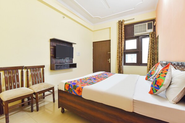 A hotel room with a bed, a TV mounted on the wall, a wooden chair, and a window with curtains.