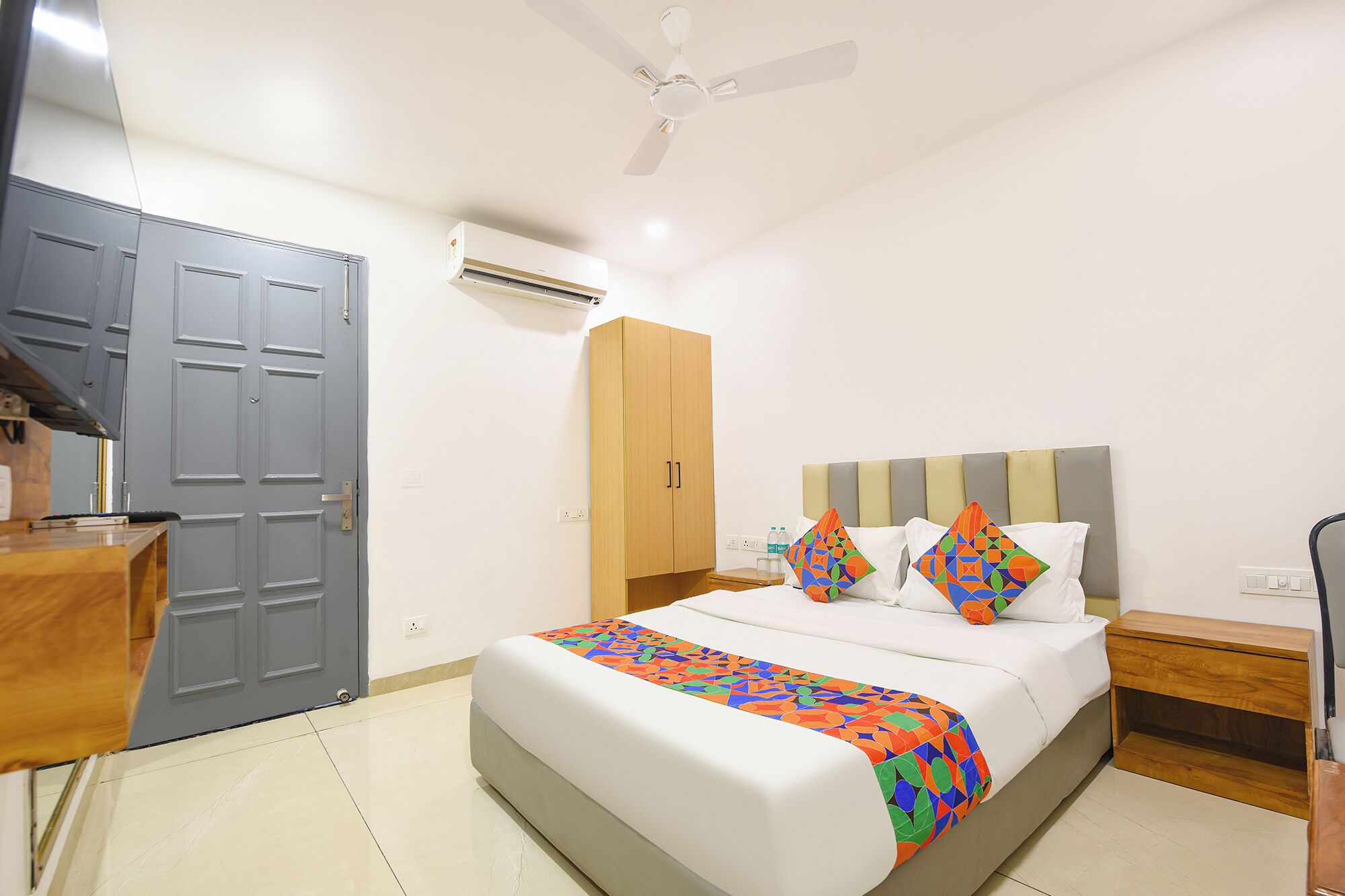 A hotel room with a bed, a desk, a wardrobe, a ceiling fan, and an air conditioning unit.