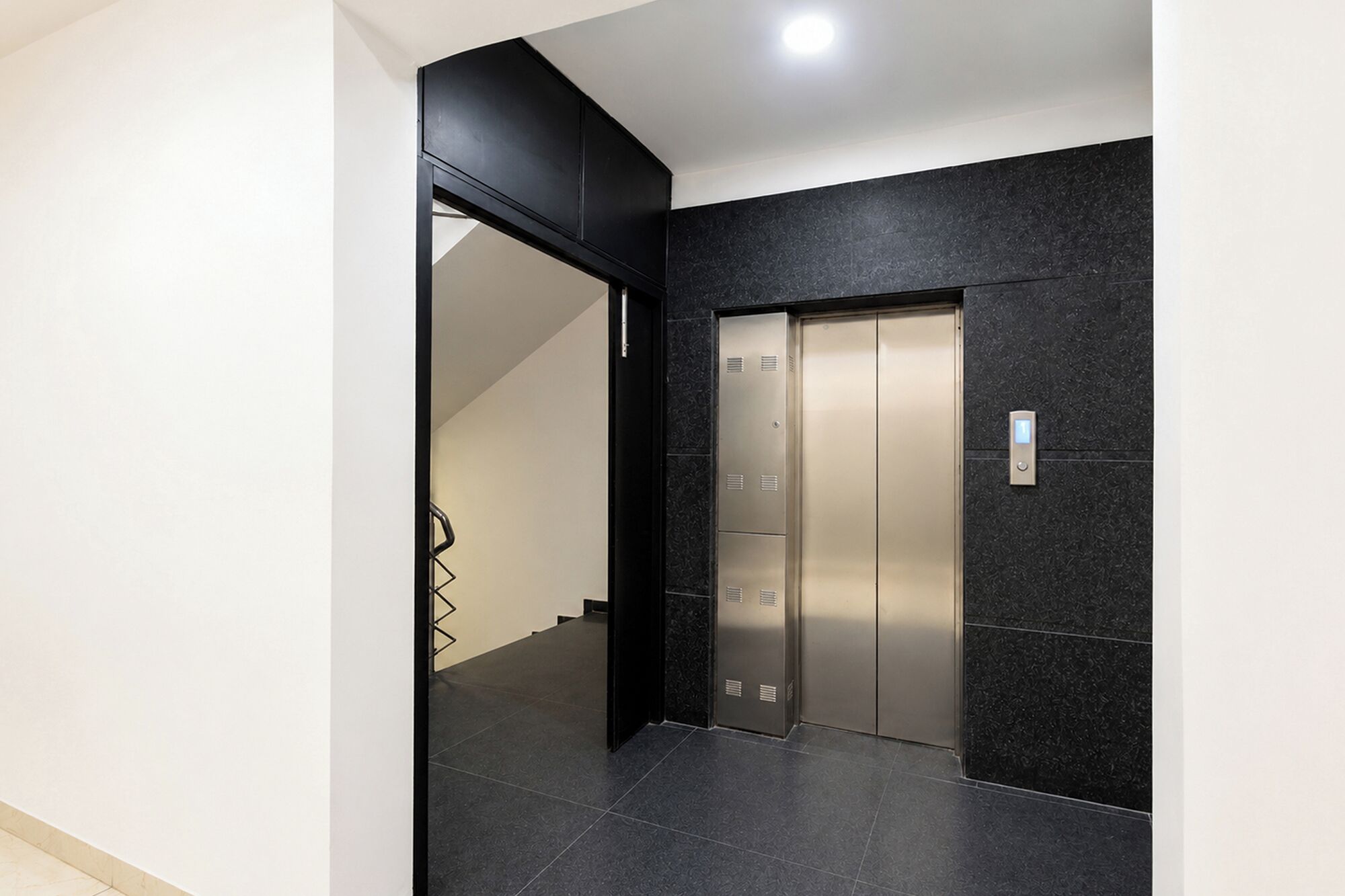 A modern elevator with a metallic finish, located in a hallway with dark tiled flooring and a black accent wall.