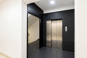 Un ascensor moderno con detalles metálicos, situado en un pasillo con suelo de baldosas oscuras y una pared de acento negra.