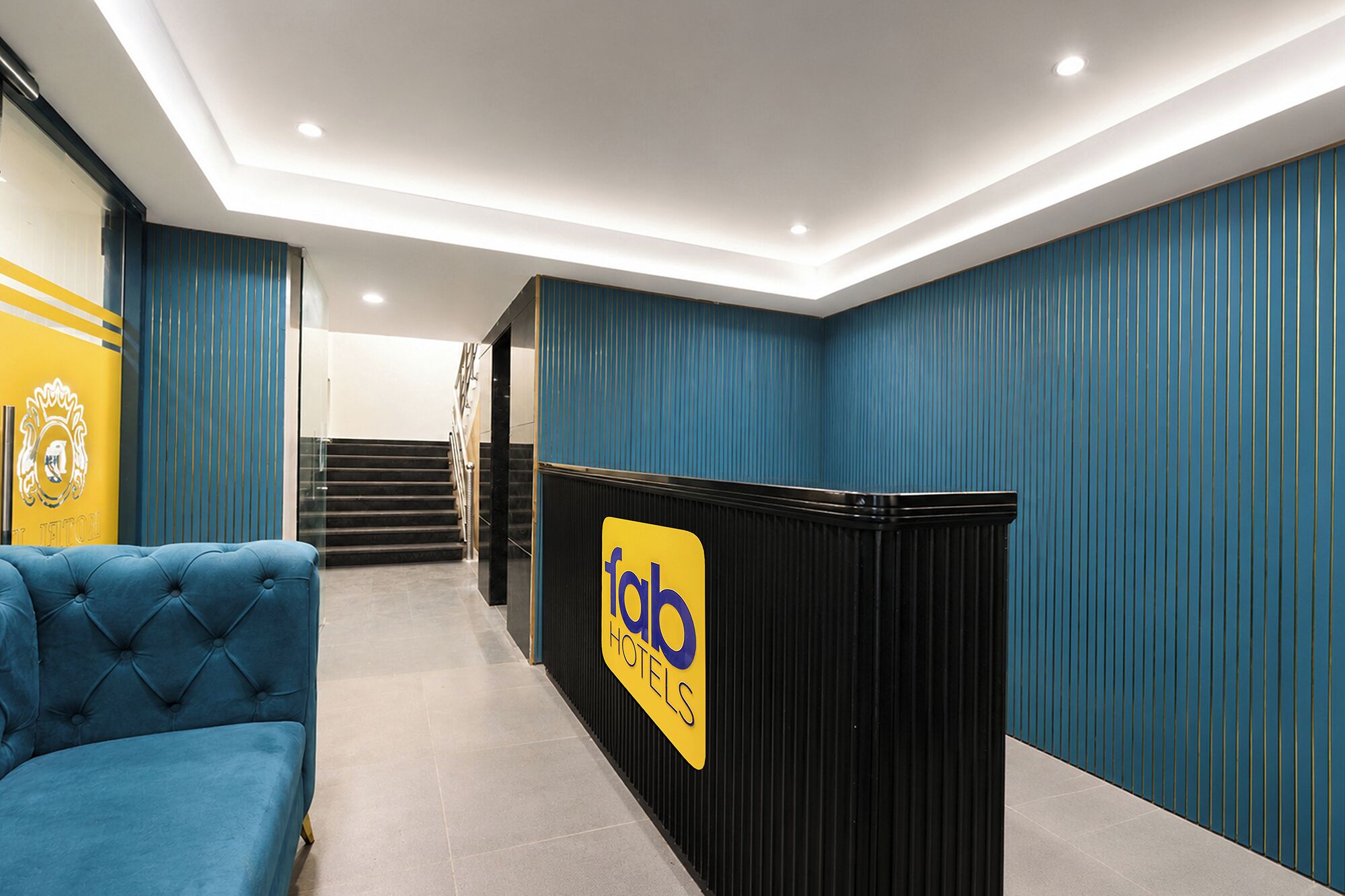 A hotel reception area with a blue upholstered sofa, a reception desk with a logo, and a staircase leading to an upper floor.