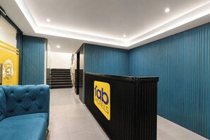 Zona de recepció d'hotel amb un sofà blau tapissat, una taula de recepció amb un logotip i una escala que condueix a un pis superior.