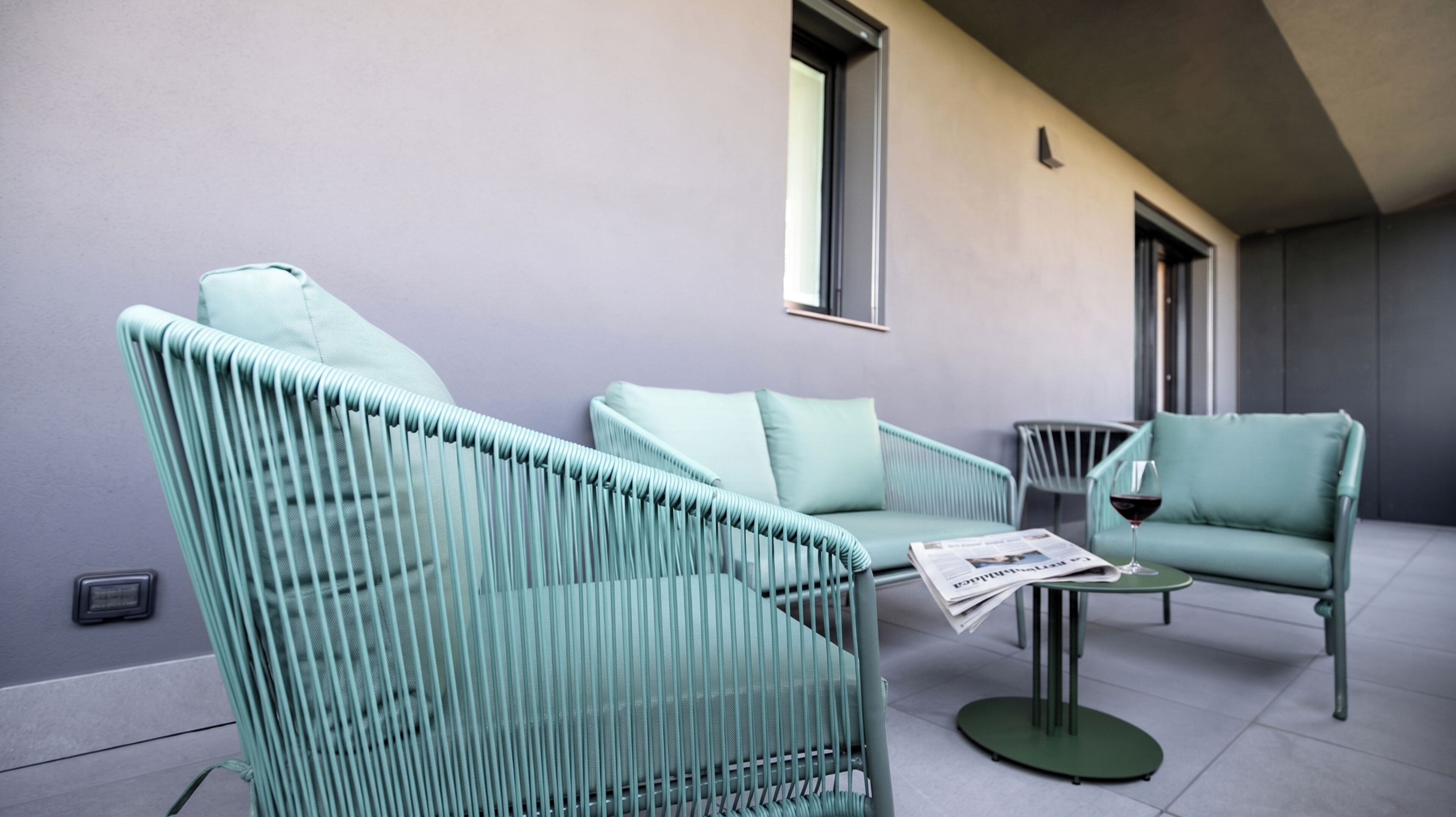 A modern outdoor seating area with light green upholstered chairs and a small table, featuring a newspaper and a glass of red wine.
