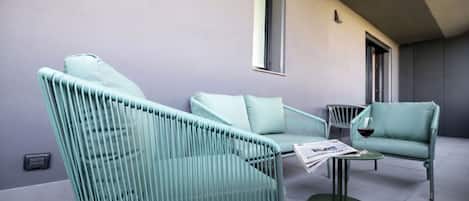 Un espace extérieur moderne, aménagé avec des chaises rembourrées de couleur vert clair et une petite table, où l’on peut trouver un journal et un verre de vin rouge.