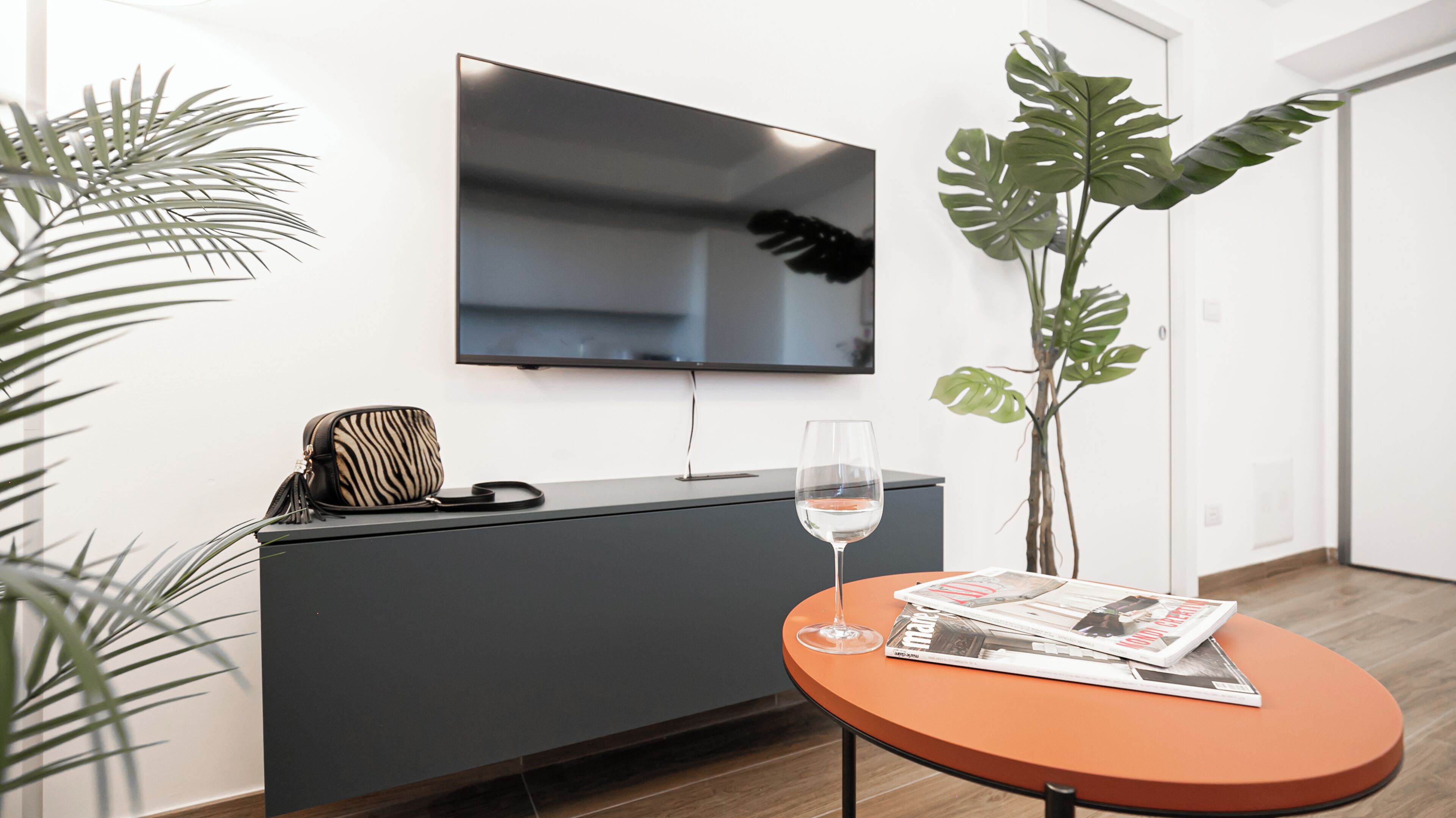 A modern living room with a flat-screen TV mounted on the wall, a grey entertainment unit, a glass wine glass on a small orange table, and a magazine.