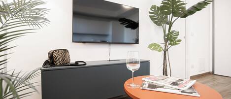 A modern living room with a flat-screen TV mounted on the wall, a grey entertainment unit, a glass wine glass on a small orange table, and a magazine.