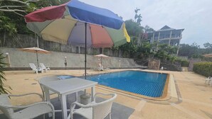 A swimming pool with lounge chairs and umbrellas, surrounded by a concrete wall and vegetation. - Chaya Village F Shared Pool & Jacuzzi (Ko Lanta)