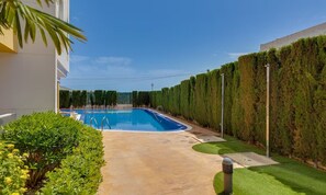 A swimming pool with a clear blue water, surrounded by a well-maintained hedge, a concrete pathway, and a small grassy area. - Apartment Santa Ana Villamartin (Orihuela Costa)