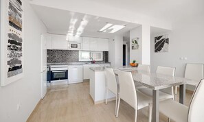 A modern kitchen with white cabinetry, a granite countertop, and a dining area with a marble table and white chairs. - Apartment Santa Ana Villamartin (Orihuela Costa)