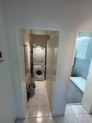 Ein kleiner Hauswirtschaftsraum mit Waschmaschine und Trockner, einer Mülltonne und einem gefliesten Boden.