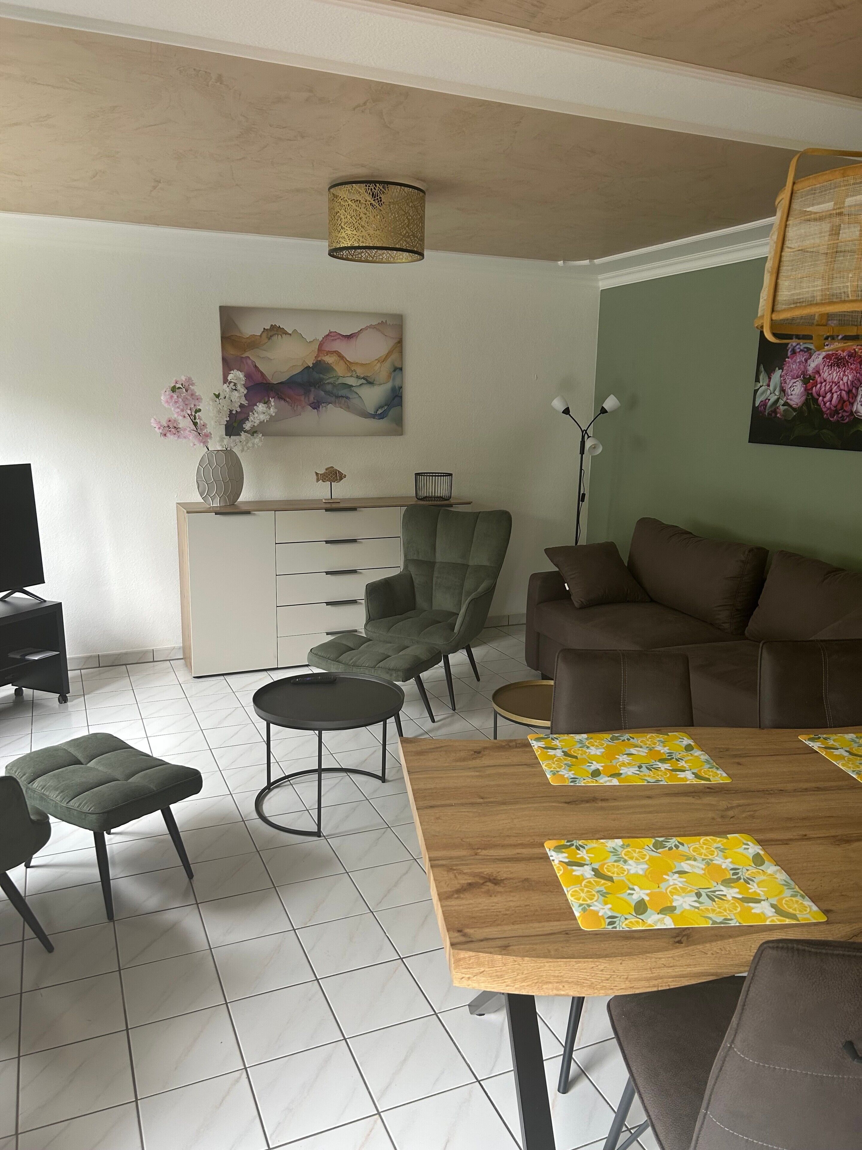 A living room with a sofa, a dining table with placemats, a green chair, a small round table, and a TV stand.