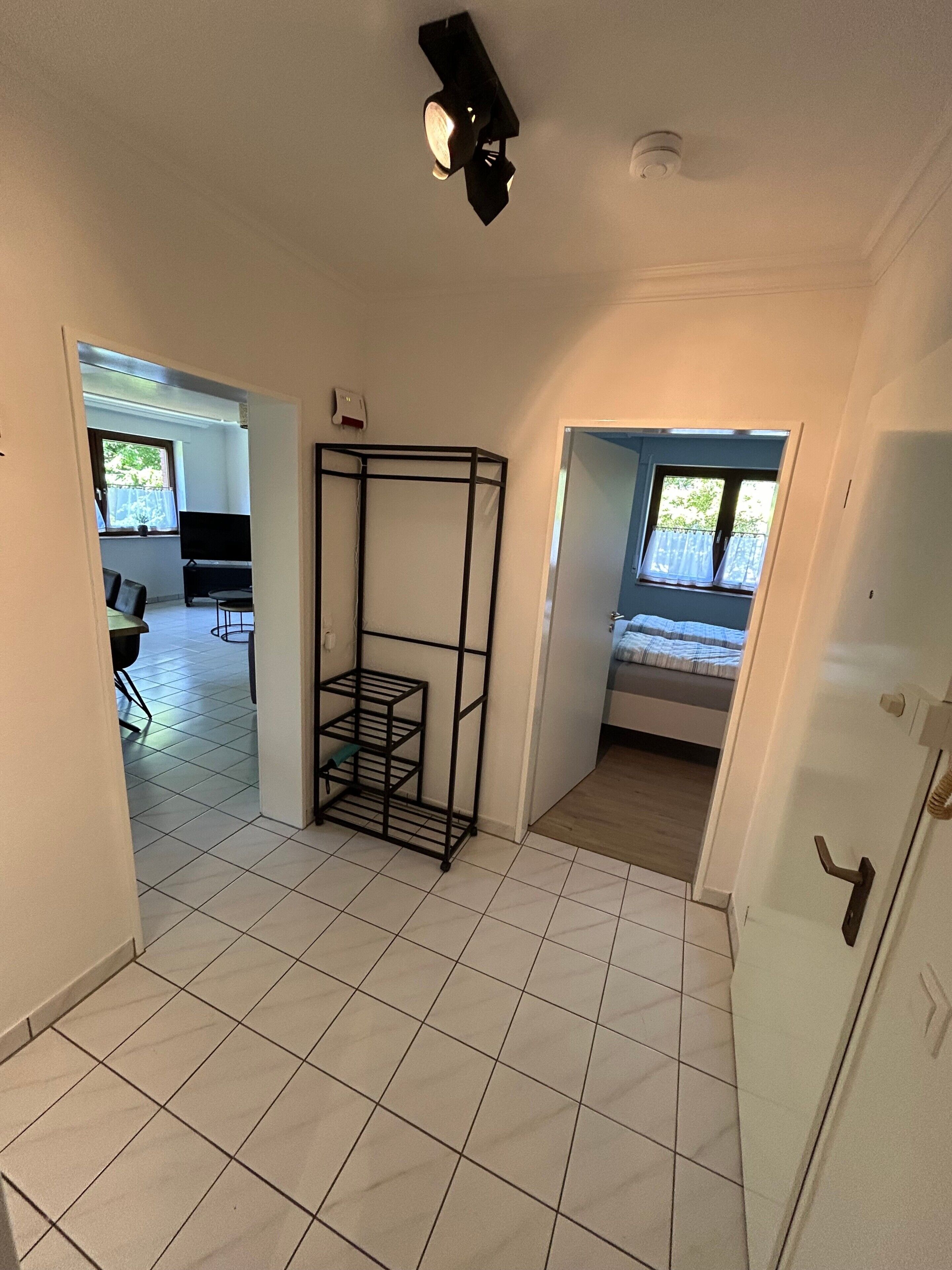A room with a tiled floor, a metal shelf unit, a bed with a window, and a door leading to another room.