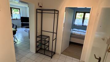 A room with a tiled floor, a metal shelf unit, a bed with a window, and a door leading to another room.