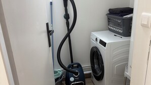 Een wasserette met een wasmachine, stofzuiger en een mand op de vloer.