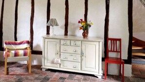 Una habitación con vigas de madera a la vista, un armario blanco con cajones, una silla roja, una silla a rayas y un florero con flores.