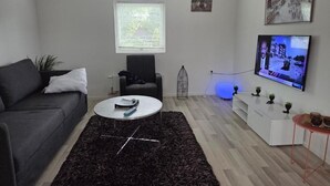 A modern living room with a grey sofa, a round coffee table, a TV mounted on the wall, a small table with decorative items, and a window with blinds. - Charming Jasmine House in Bromölla (Bromolla)