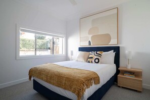 A bedroom with a bed, a nightstand, a lamp, a window, and a painting on the wall. - Unit 2 380 Drummoyne (Drummoyne)