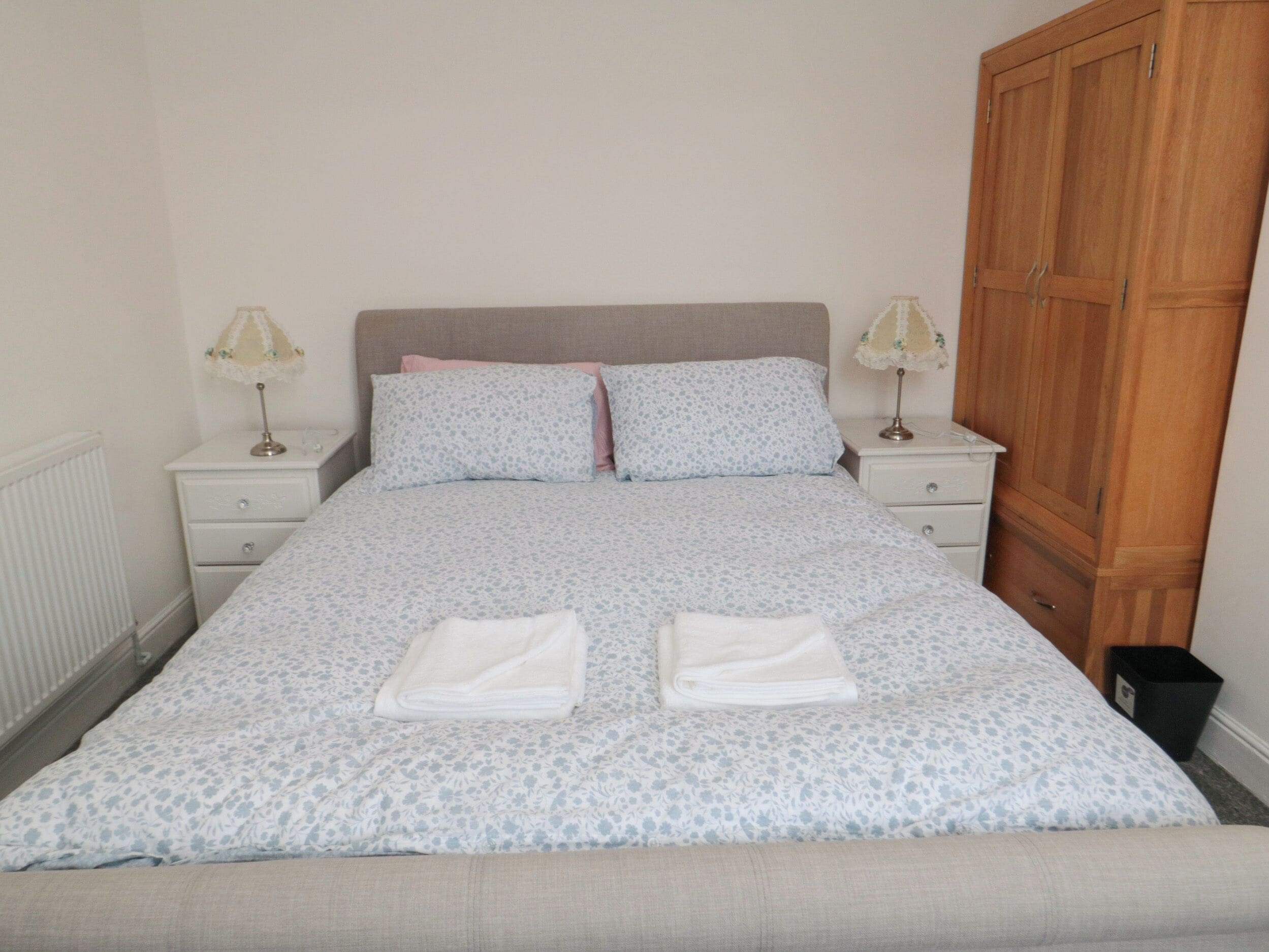 A neatly made bed with a floral-patterned bedspread, two pillows, and two folded towels. There are two bedside tables with lamps, a wooden wardrobe, and a radiator on the wall.