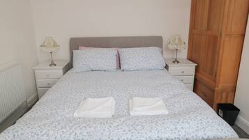 A neatly made bed with a floral-patterned bedspread, two pillows, and two folded towels. There are two bedside tables with lamps, a wooden wardrobe, and a radiator on the wall.
