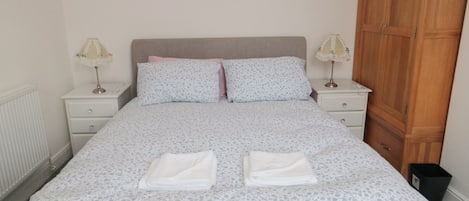 A neatly made bed with a floral-patterned bedspread, two pillows, and two folded towels. There are two bedside tables with lamps, a wooden wardrobe, and a radiator on the wall.