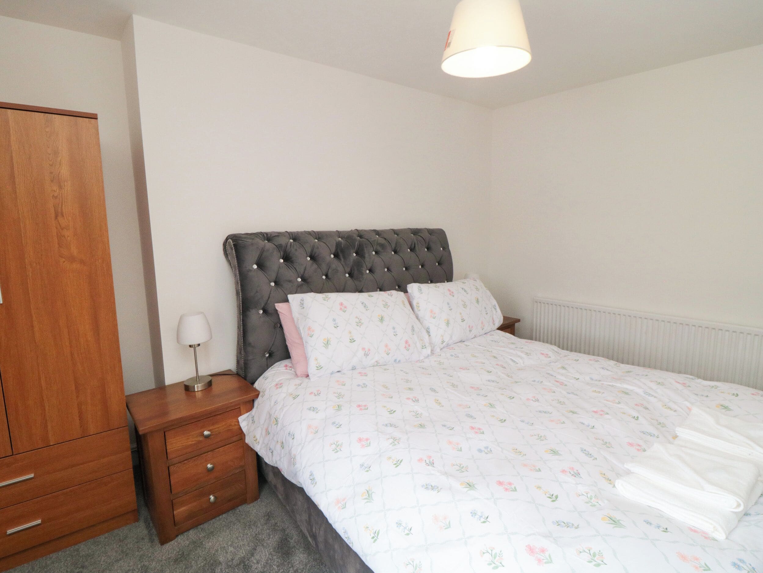 Une chambre à coucher avec une tête de lit capitonnée, une armoire en bois, une table de chevet avec une lampe et une couvre-lit à motifs floraux.