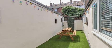 Un patio estrecho con césped artificial, un banco de madera y una pared decorada con faroles de colores.