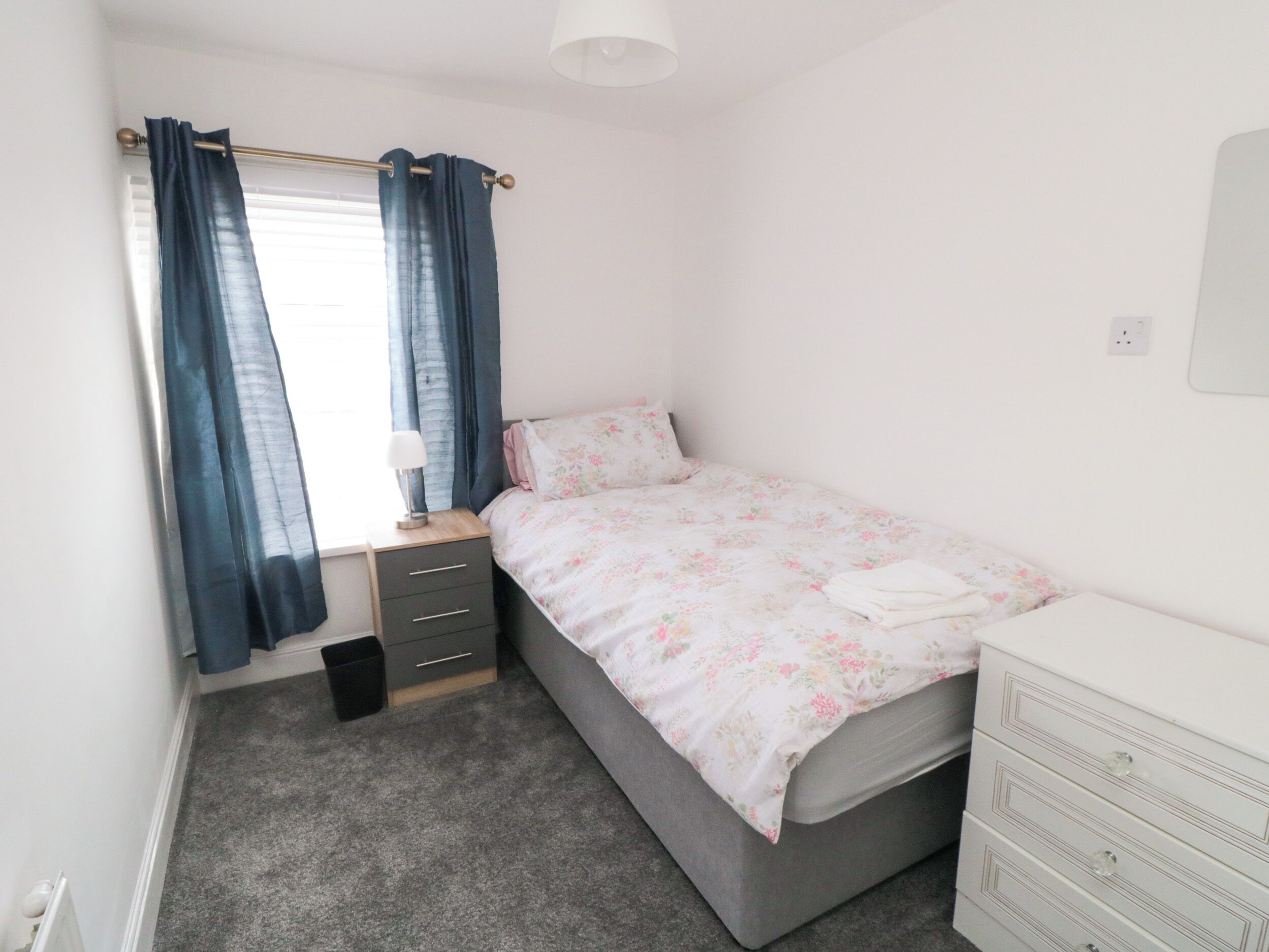 A bedroom with a single bed, a bedside table with a lamp, a dresser, and a window with curtains.