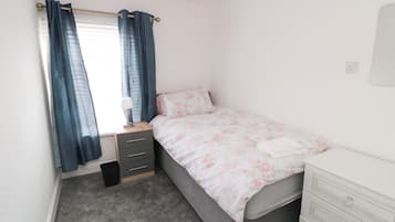 A bedroom with a single bed, a bedside table with a lamp, a dresser, and a window with curtains.