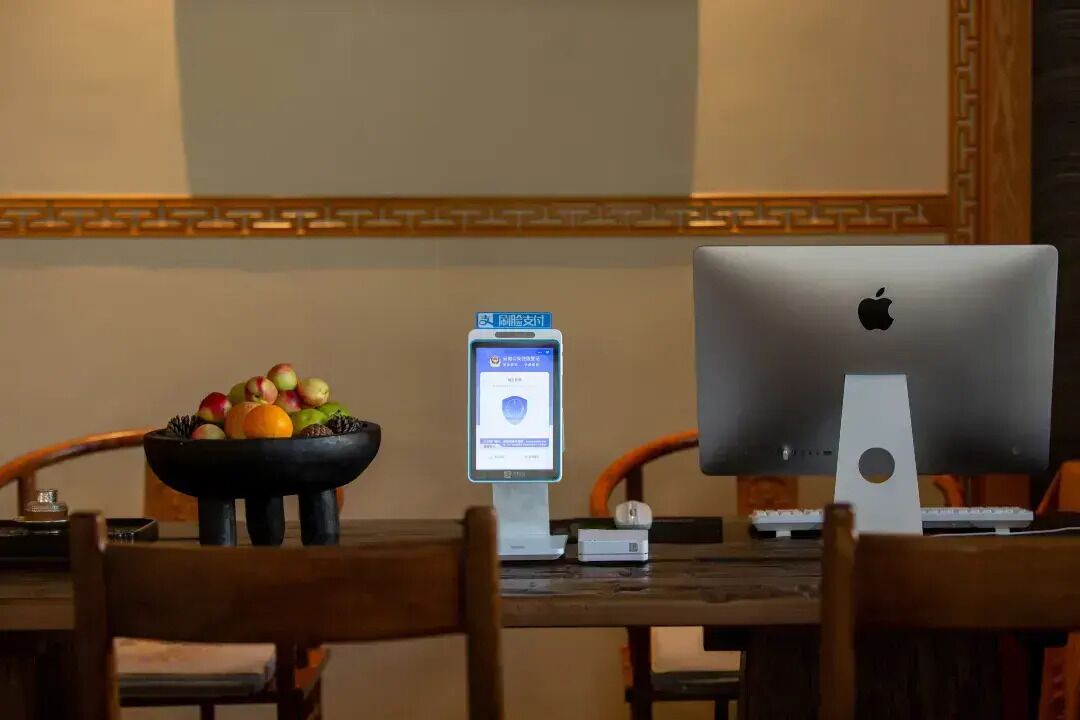 Uma mesa de jantar com um iMac da Apple, uma tigela de frutas e um pequeno dispositivo exibindo um menu.