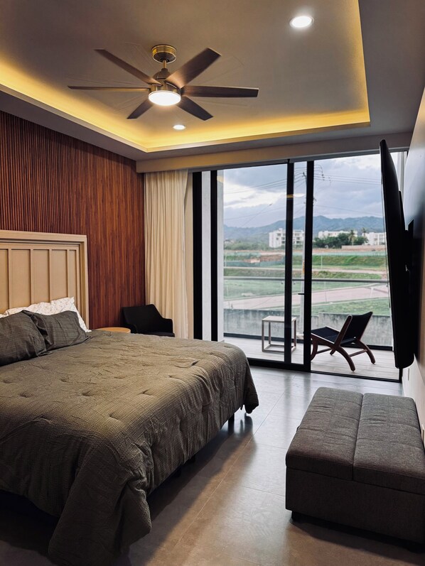 Ein modernes Schlafzimmer mit einem großen Bett, einem Deckenventilator, einem Fernseher und einem Balkon mit Blick ins Freie.