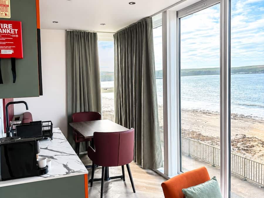 A room with a dining table, chairs, a coffee machine, a fire extinguisher, and a view of the beach.