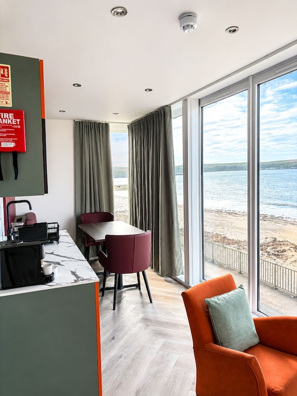 A room with a dining table, chairs, a coffee machine, a fire extinguisher, and a view of the beach.
