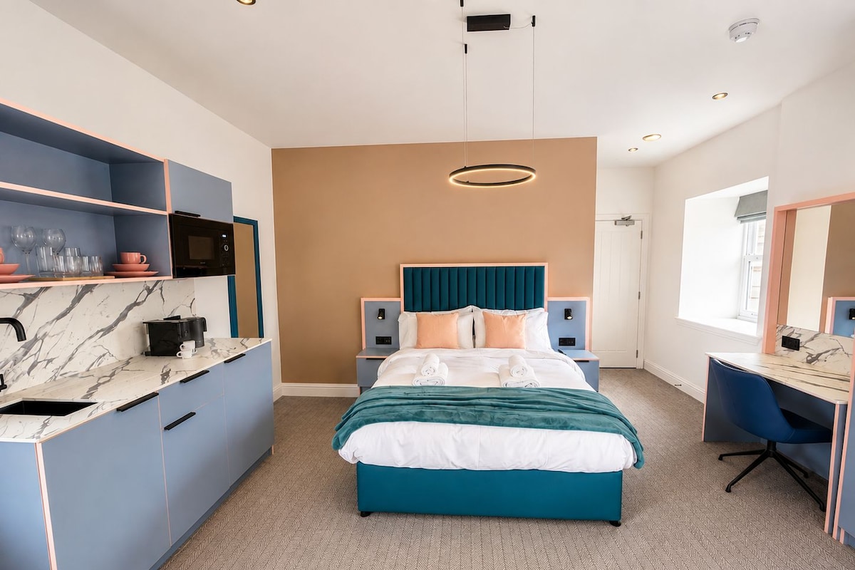 A modern hotel room with a bed, a desk, a kitchenette, and a large window.