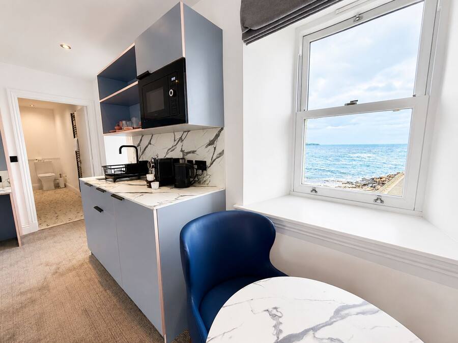 A compact kitchen with a view of the sea, a blue chair, and a marble countertop.