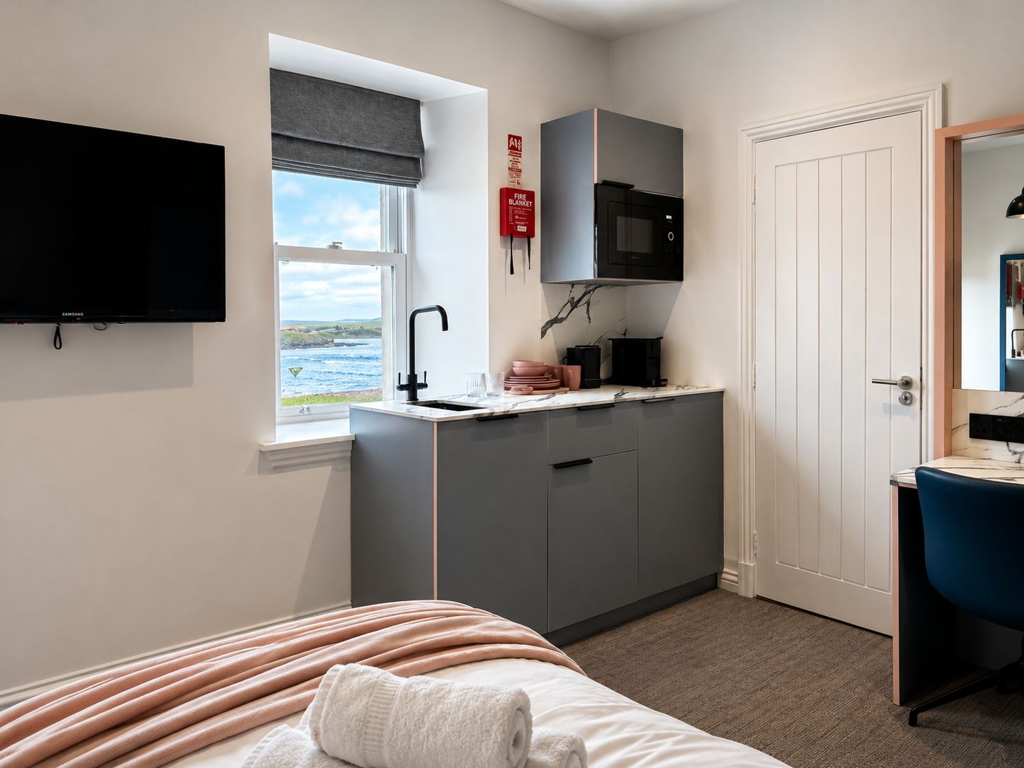 A compact hotel room with a bed, a small kitchenette, a television, and a window with a view of the sea.