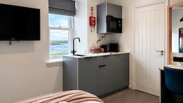 A compact hotel room with a bed, a small kitchenette, a television, and a window with a view of the sea.