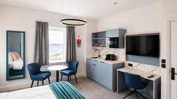A hotel room with a bed, a desk with a TV, a seating area with chairs, a window with a view of the beach, and a kitchen area with a microwave.