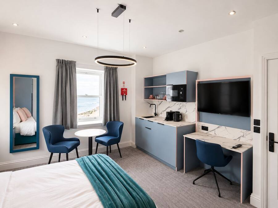 A hotel room with a bed, a desk with a TV, a seating area with chairs, a window with a view of the beach, and a kitchen area with a microwave.