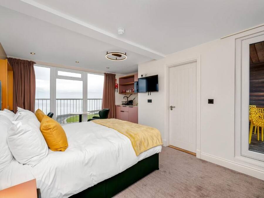 A hotel room with a bed, a desk, a television, and a balcony with yellow chairs.