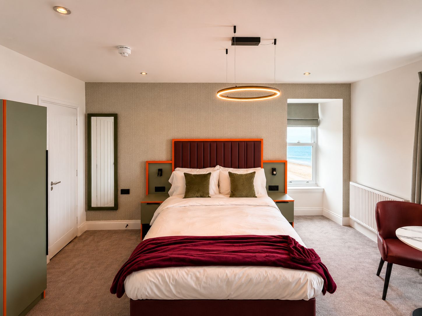 Quarto de hotel com cama, mesa de cabeceira, poltrona, janela com vista para a praia e luminária no teto.
