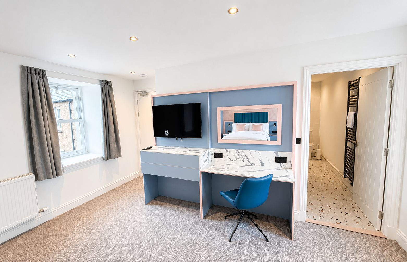 A modern bedroom with a bed, a desk, a chair, a TV, and a bathroom visible through an open door.
