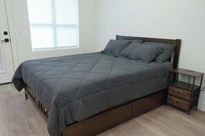 A bed with a dark gray quilt, a wooden headboard, and a nightstand with drawers. - Modern Studio Near Airport with Free Parking (Millcreek)