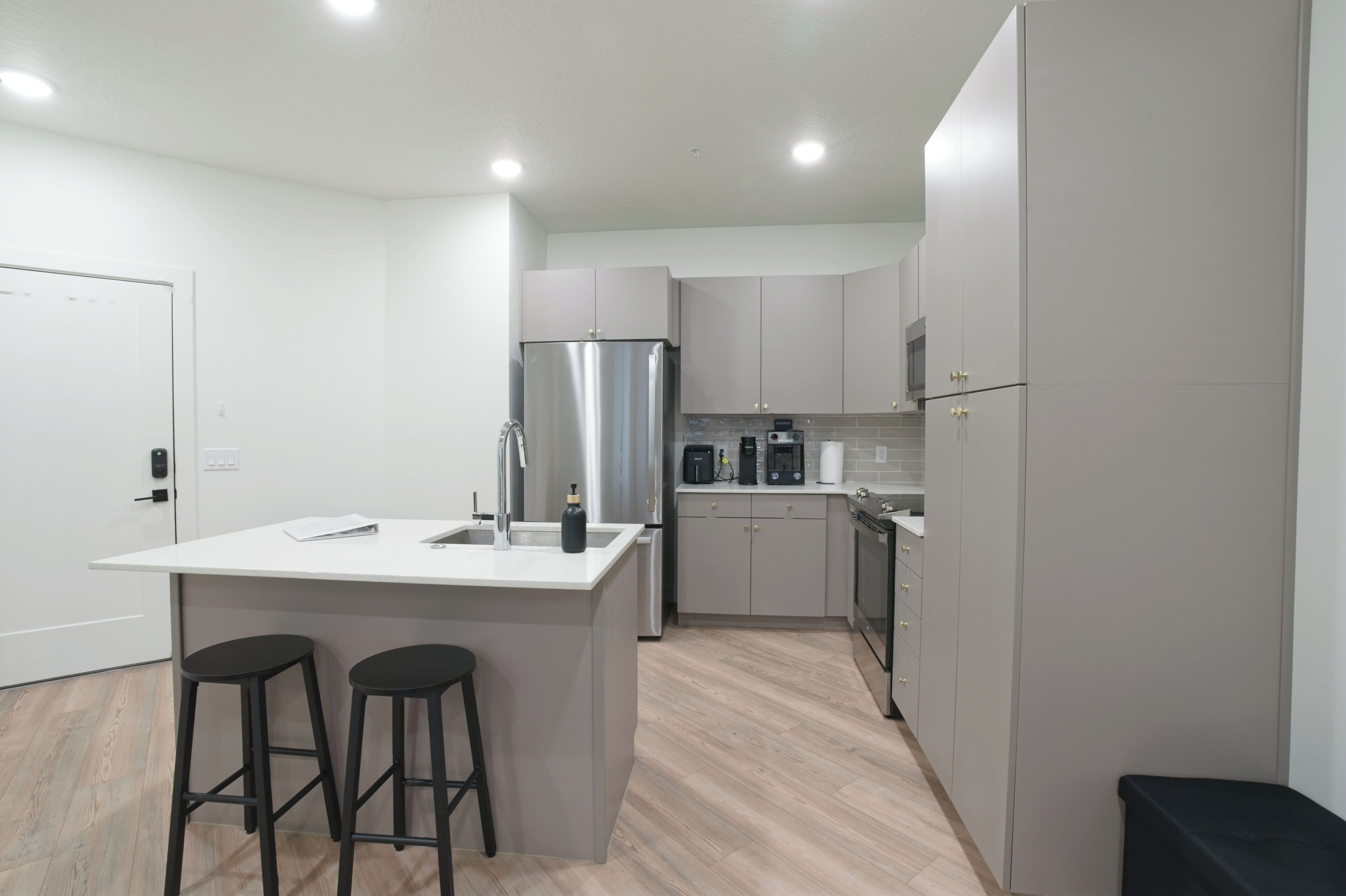 A modern kitchen with a central island, bar stools, a stainless steel refrigerator, and a coffee maker.