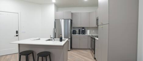 A modern kitchen with a central island, bar stools, a stainless steel refrigerator, and a coffee maker.